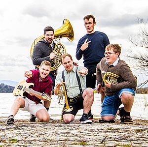 Die fünf Bandmitglieder mit ihren Blasinstrumenten vor einem See und Bergpanorama